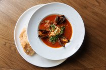 Vue du dessus de l'assiette ronde blanche avec une riche soupe de tomates épicée aux moules noires et aux fruits de mer garnis de légumes verts hachés — Photo de stock