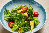De cima de salada com pedaços de tomate cereja abacate e folhas de arugula vestida com azeite em tigela azul — Fotografia de Stock