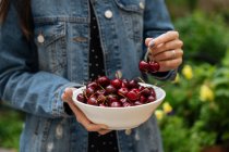 Faceless woman in jeans jacket holding white bowl with fresh cherry standing with green plants on blurred background — Stock Photo