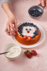 Dall'alto di donna irriconoscibile decorato torta fatta in casa con mirtillo e fragola sul tavolo con ciotola di panna acida — Foto stock