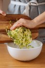 Crop housewife putting grating zucchini from wooden cutting board into large white bowl while preparing food an wooden table — Stock Photo