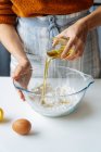 Ritaglia l'olio d'oliva dal bicchiere in una grande ciotola con la farina mentre prepari l'impasto a tavola bianca in cucina. — Foto stock