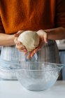 Crop housewife in casual clothes and apron holding dough in hands while standing at table with large glass bowl — Stock Photo