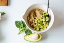 From above of white ceramic bowl with quinoa and chopped avocado placed on white table with half of avocado and green basil leaves — Stock Photo