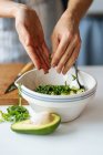 Crop hands of housewife adding chopped fresh green herbs into bowl with avocado while preparing food at white table in kitchen — Stock Photo