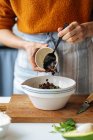 Кроп женщин добавления черного изюма в белой керамической миске помещены на деревянной доске резки во время приготовления пищи на домашней кухне — стоковое фото