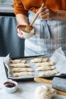 Cocinar sosteniendo el tazón y cepillando croissants en bandeja para hornear en la mesa - foto de stock