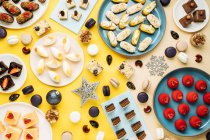 Vista superior de bugigangas de Natal e flocos de neve colocados perto de pratos com vários doces no fundo amarelo — Fotografia de Stock