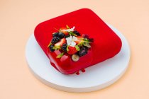 Du gâteau carré au dessus avec glaçage rouge vif et diverses baies et fleurs fraîches placées sur un plateau rond sur fond de pêche — Photo de stock