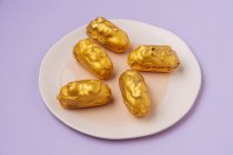Dall'alto piccoli eclairs freschi con glassa d'oro posta su piatto su sfondo lilla — Foto stock