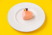 Сверху десерт в форме сердца с розовой глазурью и небольшим цветком, помещенным на тарелке на желтом фоне — стоковое фото