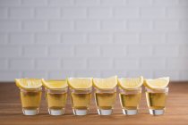 Fila di sots di vetro con tequila dorata e fette di limone su tavolo di legno con parete bianca su sfondo sfocato — Foto stock