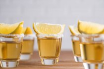 Fila de sabões de vidro com tequila dourada e fatias de limão na mesa de madeira com parede branca no fundo embaçado — Fotografia de Stock