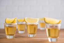 Fila de cerdas de vidrio con tequila dorado y rodajas de limón sobre mesa de madera con pared blanca sobre fondo borroso - foto de stock