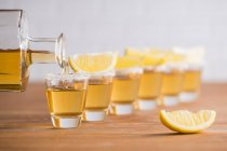 Ряд стеклянных сотов с золотой текилой и ломтиками лимона на деревянном столе с белой стеной на размытом фоне — стоковое фото