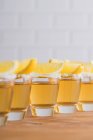 Rangée de sots en verre avec tequila dorée et tranches de citron sur table en bois avec mur blanc sur fond flou — Photo de stock