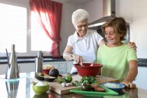 Cheerful old woman with white hair helping child while preparing guacamole together in kitchen — Stock Photo