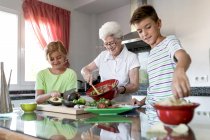 Mulher idosa alegre com cabelos brancos ajudando crianças enquanto prepara guacamole juntos na cozinha — Fotografia de Stock
