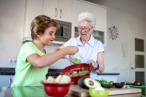 Cheerful grandma and boy tasting homemade guacamole paste with tortilla chips in kitchen — Stock Photo