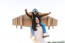 Happy cute boy wearing goggles and cardboard wings while sitting on mother shoulders and imitating aviator on meadow in backlit — Stock Photo