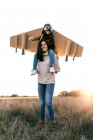 Счастливый милый мальчик в очках и картонных крыльях, сидя на плечах матери и имитируя летчика на лугу в подсветке — стоковое фото