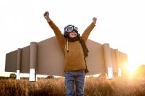 Щаслива дитина в окулярах і картонних крилах піднімає руки під час гри на полі під підсвічуванням — стокове фото