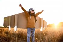Happy kid in goggles and cardboard wings raising hands during game on field in backlit — Stock Photo