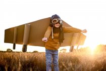 Счастливый ребенок в очках и картонных крыльях поднимает руки во время игры на поле в подсветке — стоковое фото