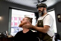 Side view of barber doing face massage to handsome redhead man with sitting in chair — Stock Photo