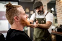 Barber with trimmer cutting beard of redhead man sitting in barbershop — Stock Photo
