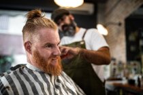 Peluquería moderna peluquería cortar el pelo de un hombre adulto pelirroja en silla de barbero - foto de stock