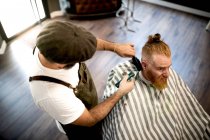 De arriba peluquero moderno peluquero cortar el pelo de un hombre adulto pelirroja en silla de barbero - foto de stock