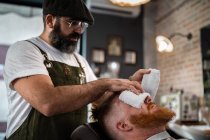 Barber doing face massage to handsome redhead man with closed eyes sitting in chair with warm hot towel covering face — Stock Photo