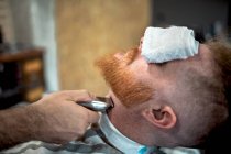 Recadré barbier méconnaissable avec tondeuse coupe rousse homme barbe avec serviette couvrant les yeux assis dans le salon de coiffure — Photo de stock