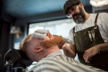 Peluquero con peine y trimmer corte pelirrojo barba hombre con toalla que cubre los ojos sentado en la barbería - foto de stock