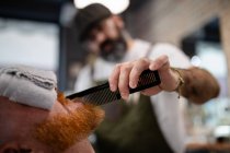 Blurred unrecognizable barber with comb fixing redhead man beard with towel covering eyes sitting in barbershop — Stock Photo