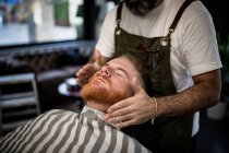 Обрезанный неузнаваемый парикмахер делает массаж лица красивому рыжему мужчине с закрытыми глазами, сидящему в кресле — стоковое фото