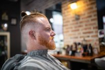 Вид сбоку на рыжего человека, сидящего в современной парикмахерской в ожидании парикмахера — стоковое фото