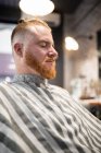 Redhead man sitting in modern barbershop with closed eyes waiting for barber — Stock Photo