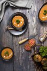 Vue du dessus des portions de purée de soupe de tomates orange dans des bols noirs élégants sur une table en bois dur avec des tranches de tomates — Photo de stock