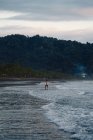 Hombre irreconocible con tabla de surf caminando cerca del mar ondeando en la noche nublada en la playa en Costa Rica - foto de stock