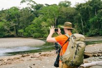 Back view of anonymous male tourist with backpack taking photos on cellphone while standing on shore in Costa Rica — Stock Photo