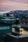High angle of solitary pickup truck with luminous headlights on wet asphalt road near cottages and industrial buildings in town against snowy mountain ridge at horizon during sunset — Stock Photo