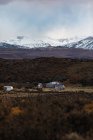 Cenário incrível com casas de campo solitárias no sopé contra topos de montanha nevados no horizonte sob céu nublado — Fotografia de Stock