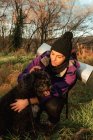 Щаслива молода жінка з рюкзаком пестить чорного собаку під час походу в сільській місцевості восени — стокове фото