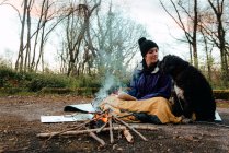 Pleased young female hiker looking at black dog while warming together at campfire in forest in autumn day — Stock Photo
