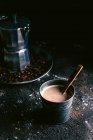 Металлическая чашка со свежеваренным кофе и деревянной ложкой на грязной черной поверхности возле подноса с кофеваркой и жареным зерном — стоковое фото
