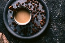 Вид зверху на кавові зерна і чашку зі свіжою кавою і ложкою, розміщені на металевому лотку на грубому столі — стокове фото