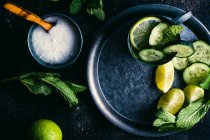 Top view of cup of sugar with spoon placed on dark table near tray with mug of fresh cucumber detox drink with mint and lime — Stock Photo