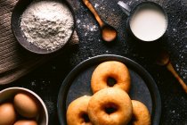 Вид зверху на свіжі пончики, розміщені на грубому столі біля різних інгредієнтів випічки та посуду на кухні — стокове фото